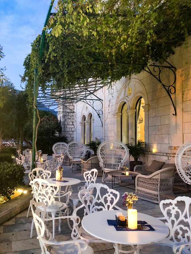 The terrace features several small white iron tables and matching chairs. A soft light glows from the rectangular candles placed on each table. An iron iron is mounted to the stone wall of the building with bright green vines dangling down through the bars.