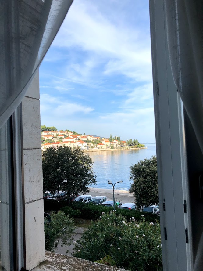 Viewing looking out a guest room window. Directly in front is a row of cars, and just beyond that is the sea, in the distance is another part of the island, lined with homes near the sea