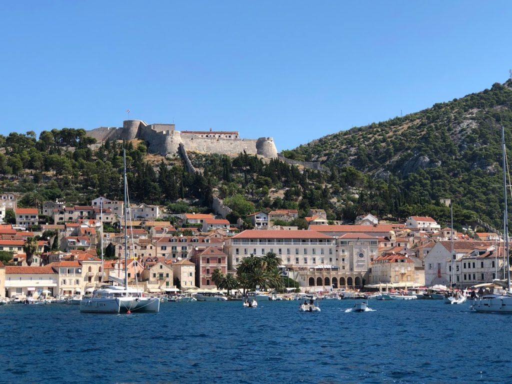 View from the harbor looking at the center of Hvar. The Palace Elizabeth sits in the center of the frame, just above the water line. In the background is the hillside and fortress 