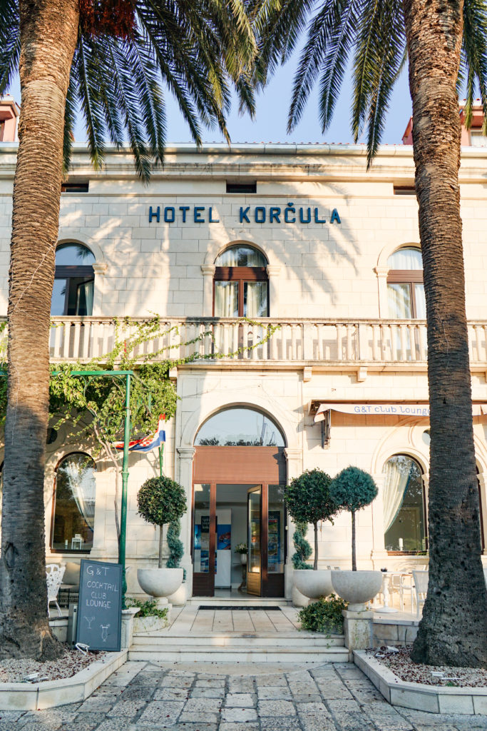 The entry is framed by two tall palms. The building is classic Dalmatian stone, the main entry door and windows are square, topped with an arch glass ornament.