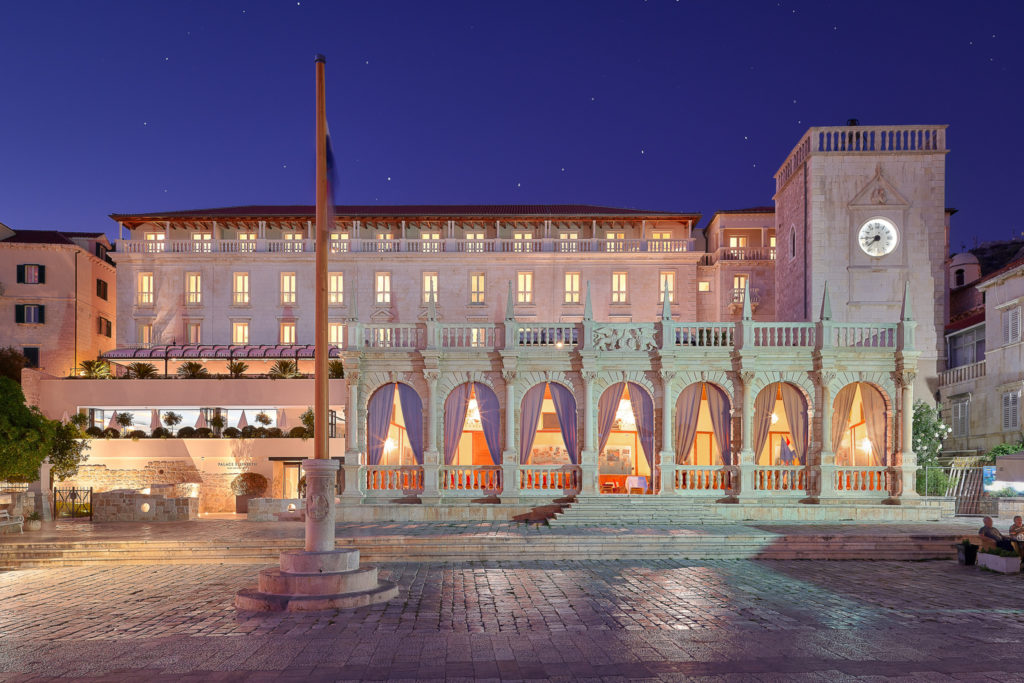 Exterior view of the hotel at night. The bright white stone façade contrasts with the clear purple sky