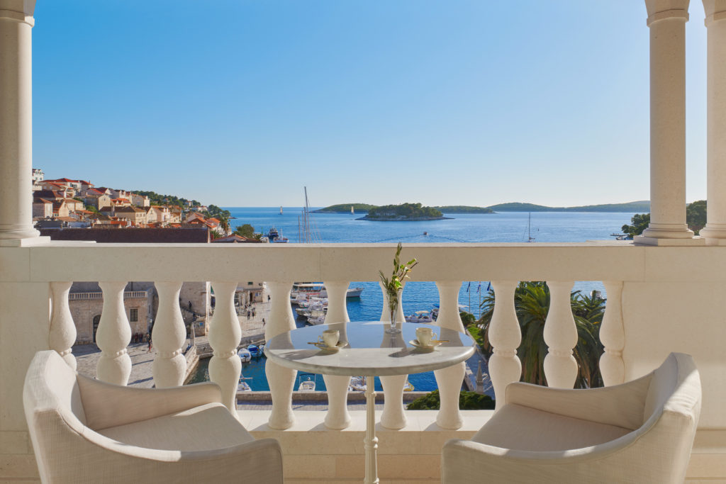 A stone patio framed by elegant columns looks out onto the harbor and the bright blue sea from a few stories up