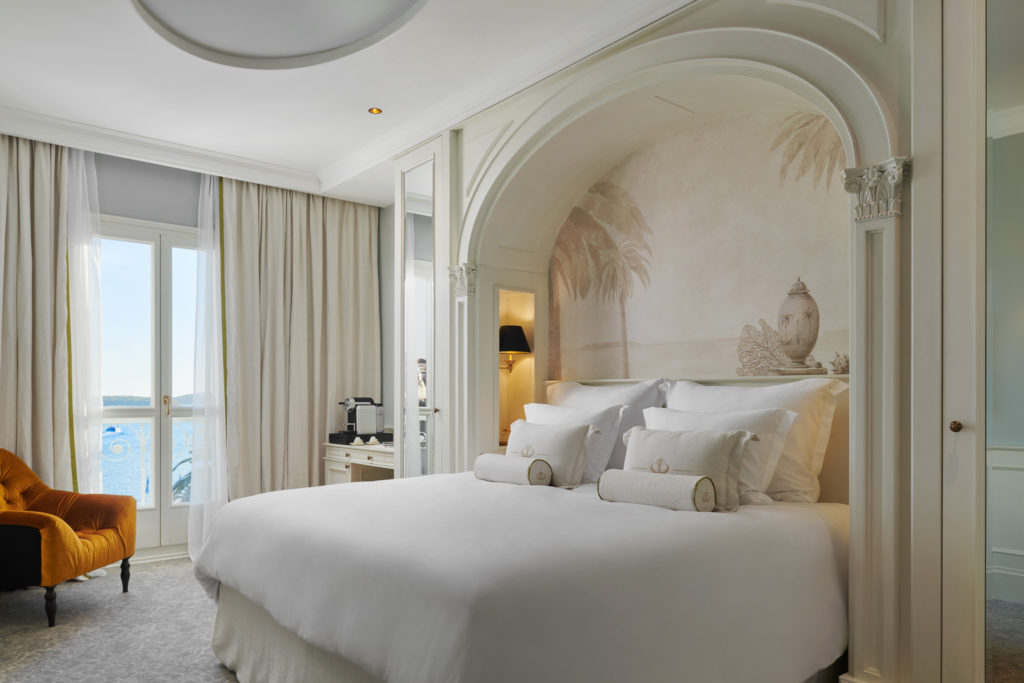 Opulent bedroom room in shades of white and cream, with a few touches of gold. Think bedding and layers of pillows in all white. The patio appears to open to view of the sea
