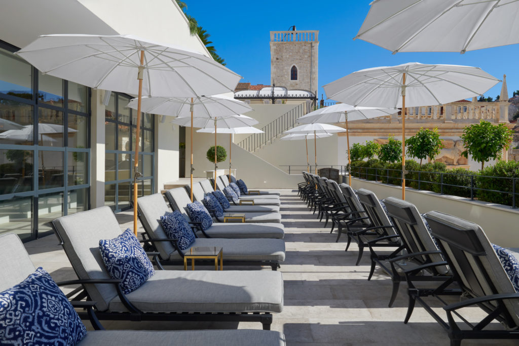 Patio chairs with plush soft grey cushions lined in row under white umbrellas. The sky above is a bright blue