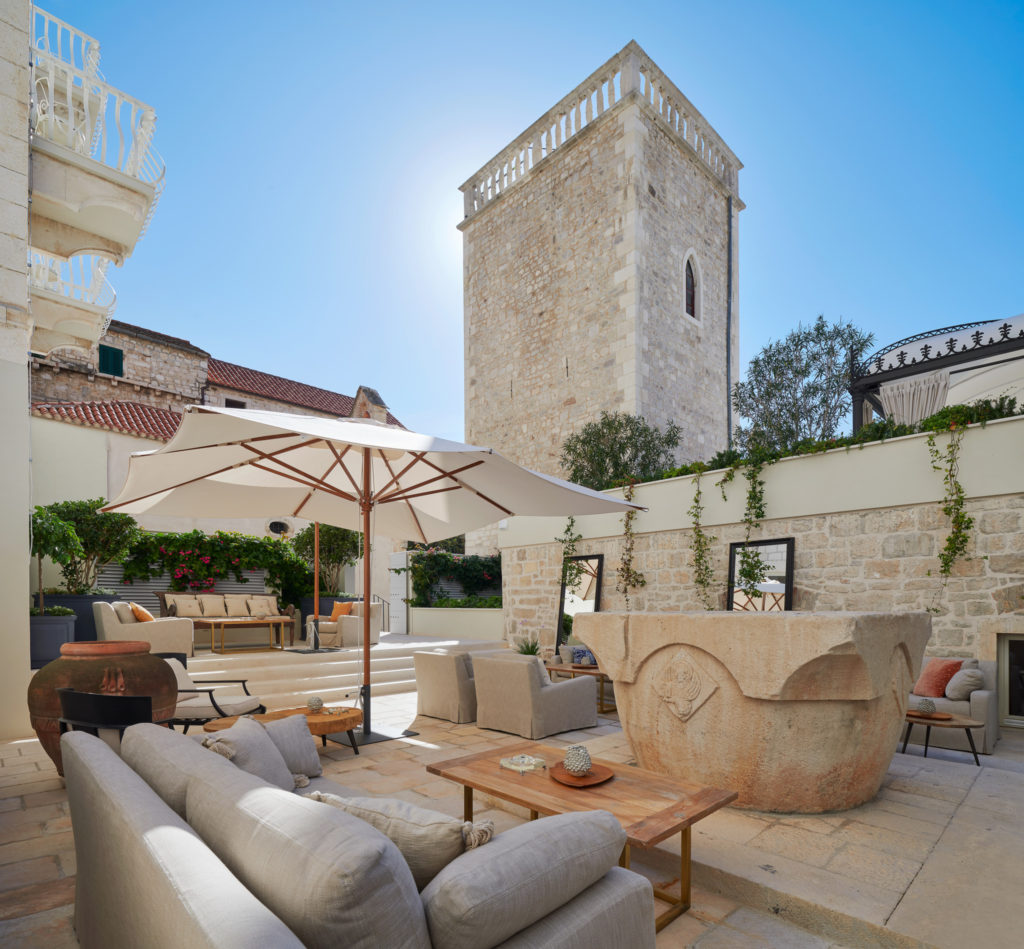 Under a bright blue sky, the stone courtyard is furnished with oversized chairs, wood tables, white sun umbrellas and a large above ground stone fountain