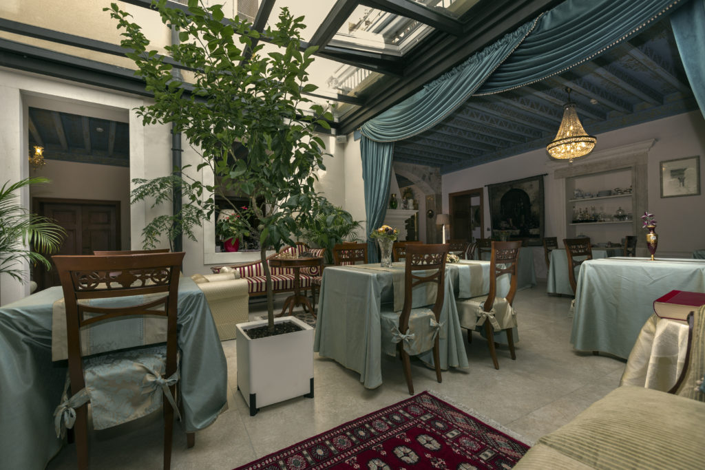 Indoor patio dining area with elegant tables dressed in pale blue and gold clothes. The ceiling is glass window with think black frame.