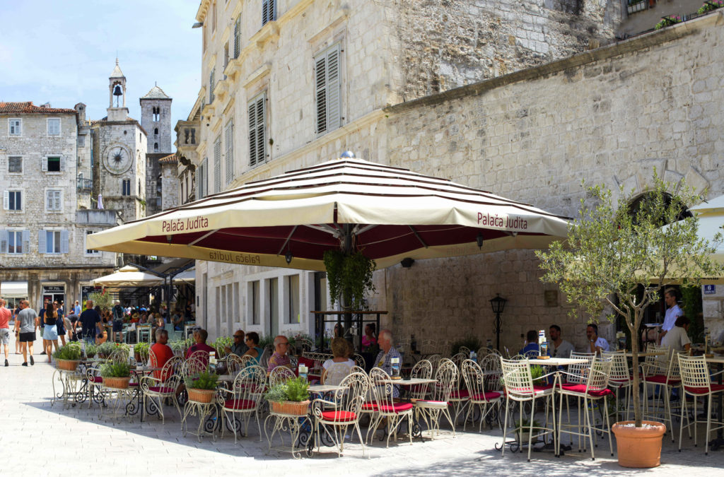 Outdoor tables on sit in front of the Judita Palace under a large pale yellow umbrella