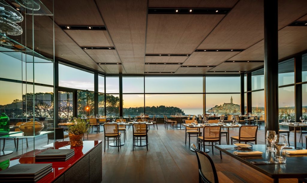 Floor to ceiling windows on three sides of the dining room of a restaurant at the Grand Park Hotel Rovinj reveal a stunning view of Rovinj and the nearby island. The clean lines of the room, and décor with minimal furnishings allow the view to be the main focal point 