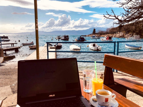 A black laptop sits on a table next to glasses of water, orange juice and coffee. In the background just a few feet beyond the table ids the sea, will several boats, and islands in the distance.