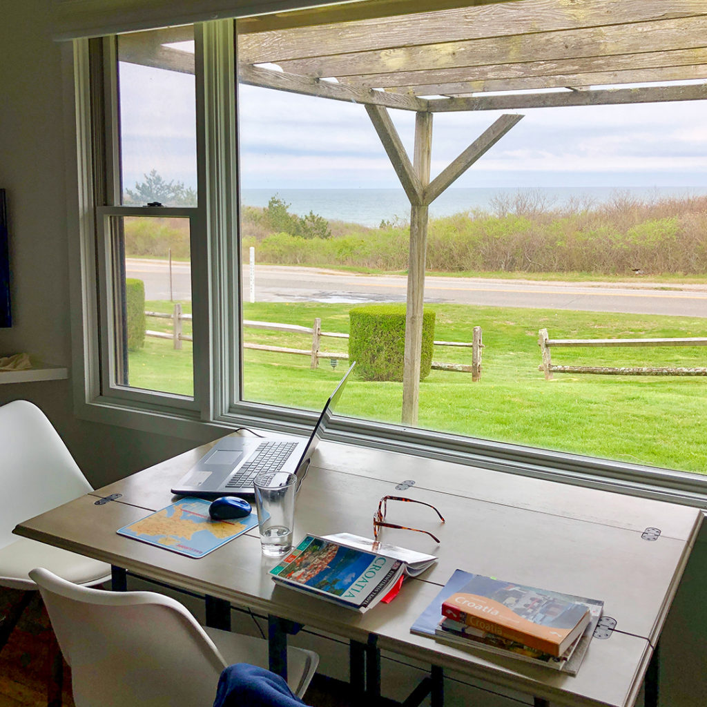 A computer sits open on a table next to a large window. In this distance the ocean can be seen